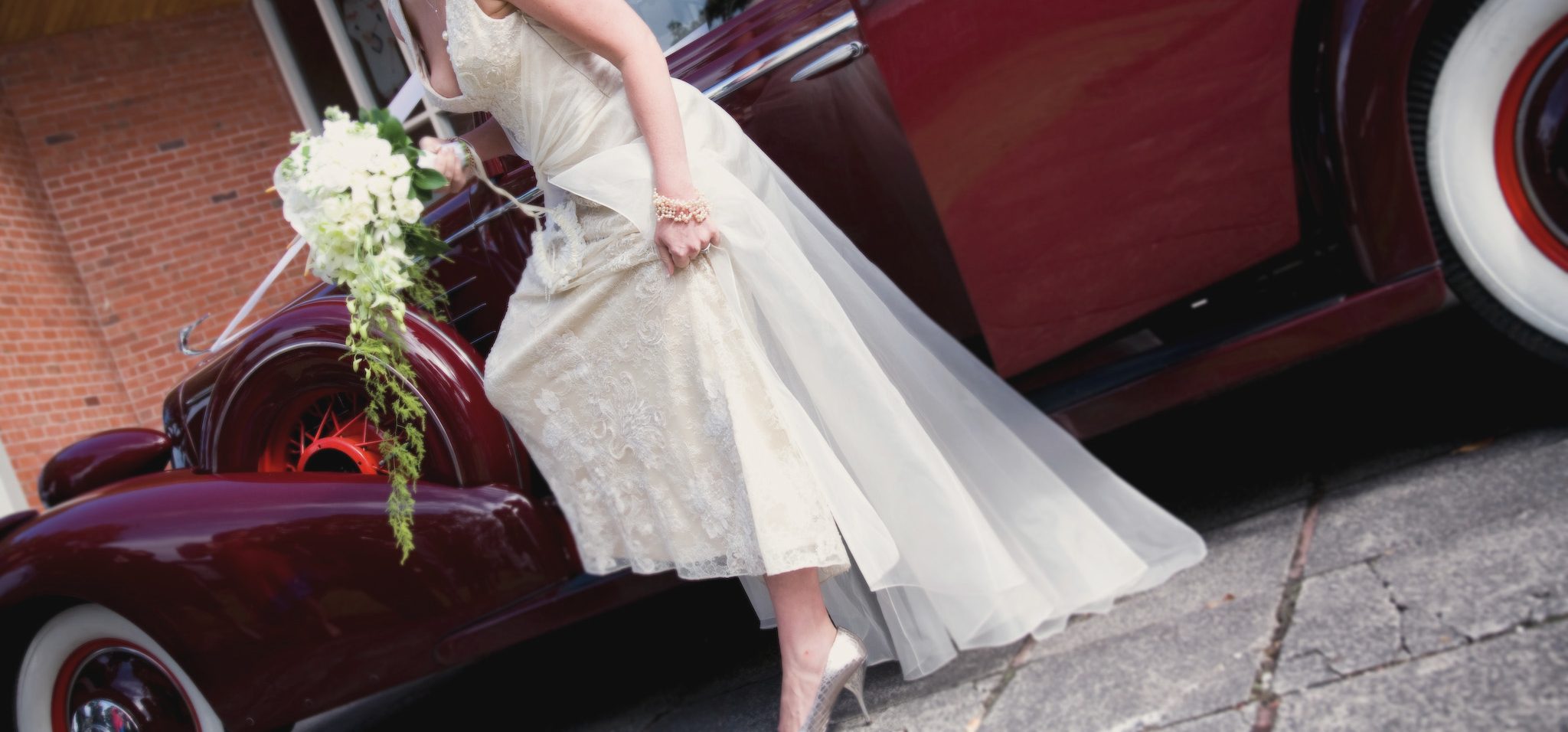 Lace wedding dress and white bouquet beside a vintage burgundy car